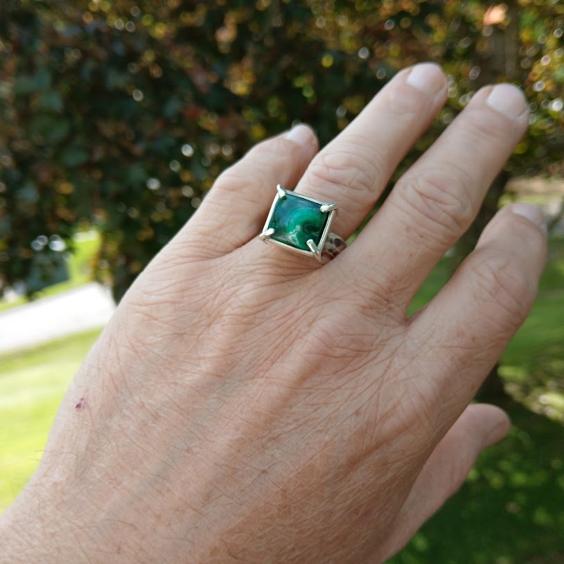 Hand wearing a ring with a green gemstone against a blurred natural background