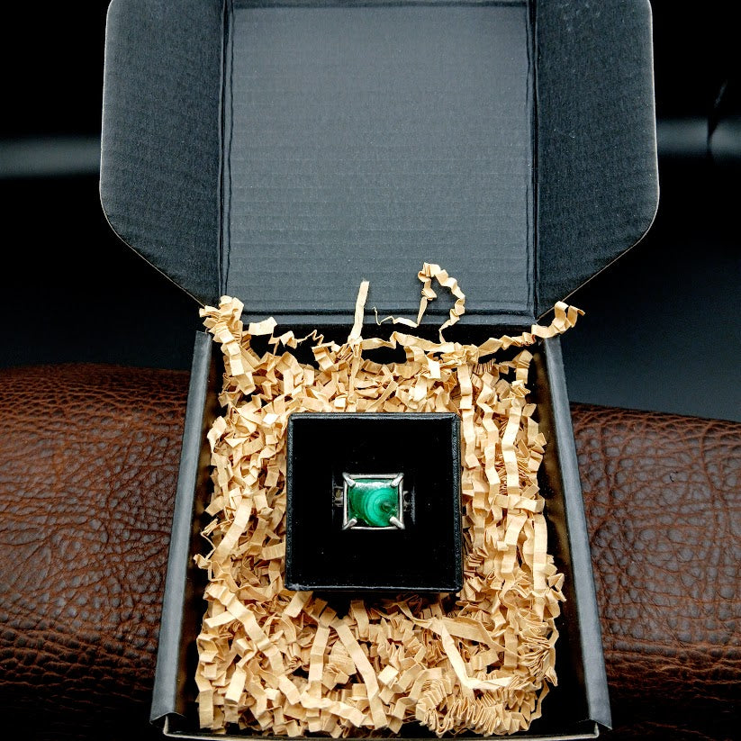 Green stone ring in black mailer box with brown crinkle cut paper around ring box