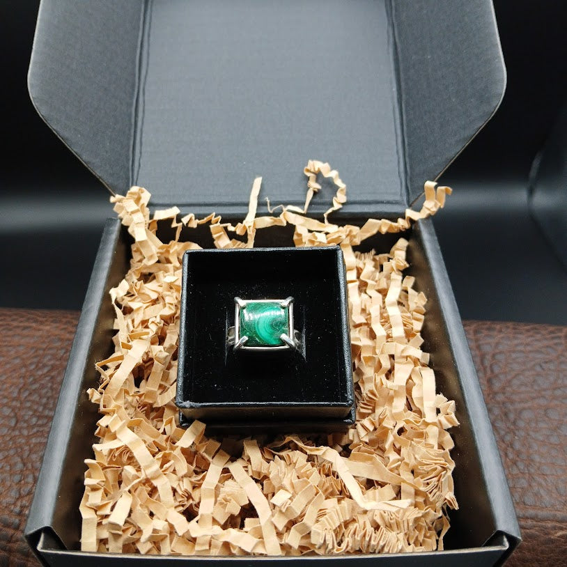 Ring with a green gemstone in a black box on crinkled paper inside an open jewelry box.