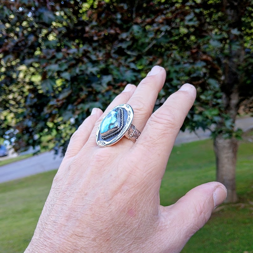 Hand wearing a ring with a turquoise gemstone against a blurred outdoor background