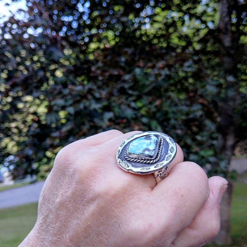 Hand wearing a silver ring with a blue stone against a blurred green outdoor background