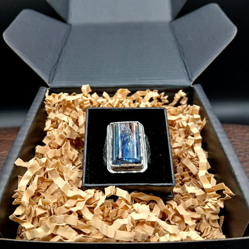 Ring with a blue gemstone in a black box on top of shredded brown paper inside an open gray box.
