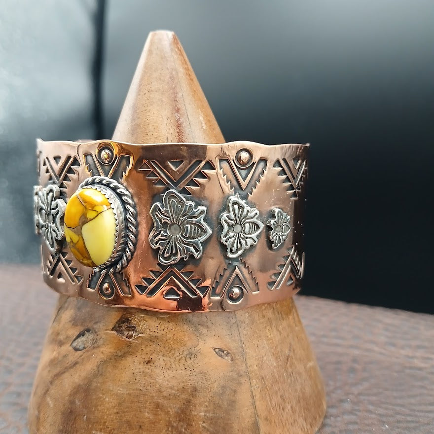 Copper bracelet with yellow stone and floral designs on a wooden stand.