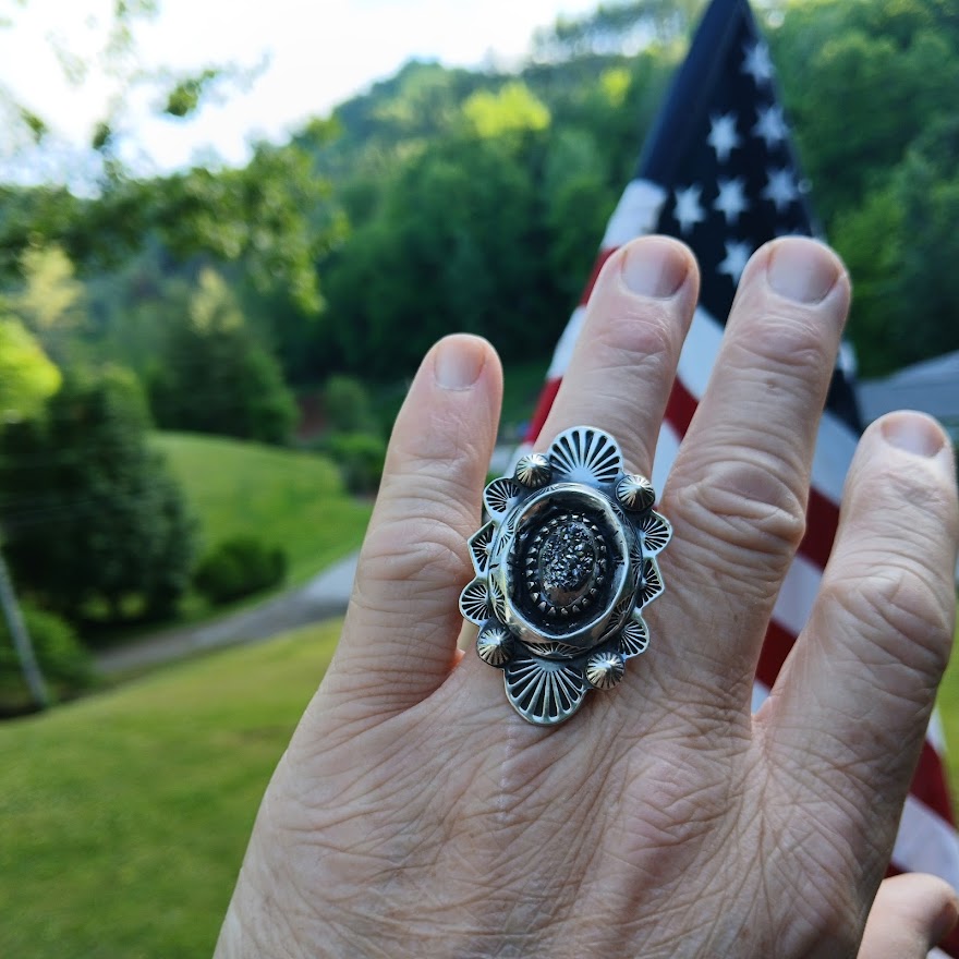 Hand wearing a silver ring with a Druzy stone center against a blurred natural background