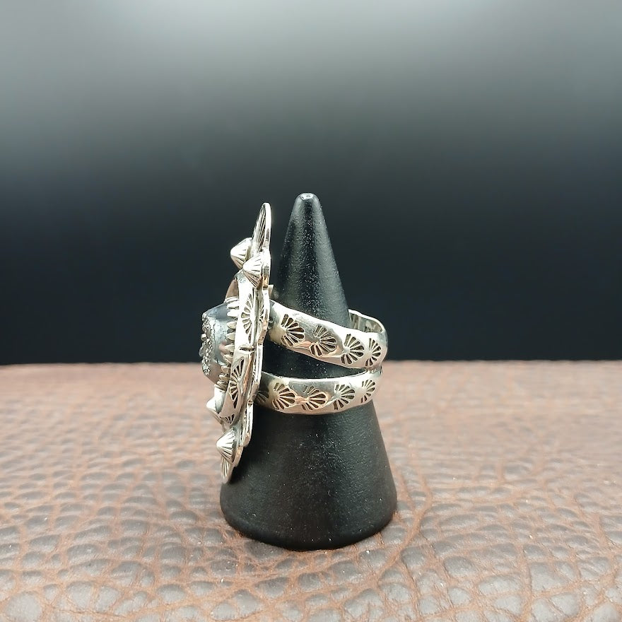 Silver ring with intricate designs on a black stand against a dark background