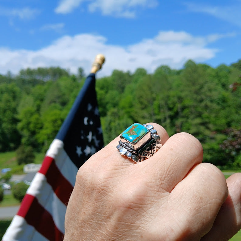 Hand wearing a ring with a turquoise stone in front of an American flag and green trees.