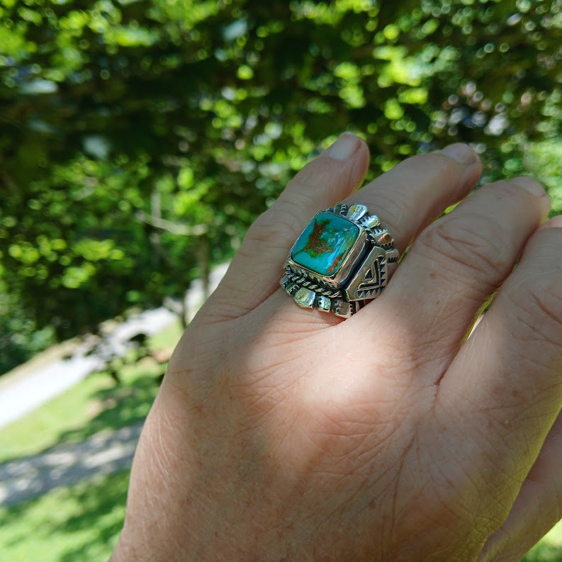 Hand wearing a large, ornate ring with a turquoise stone against a green outdoor background